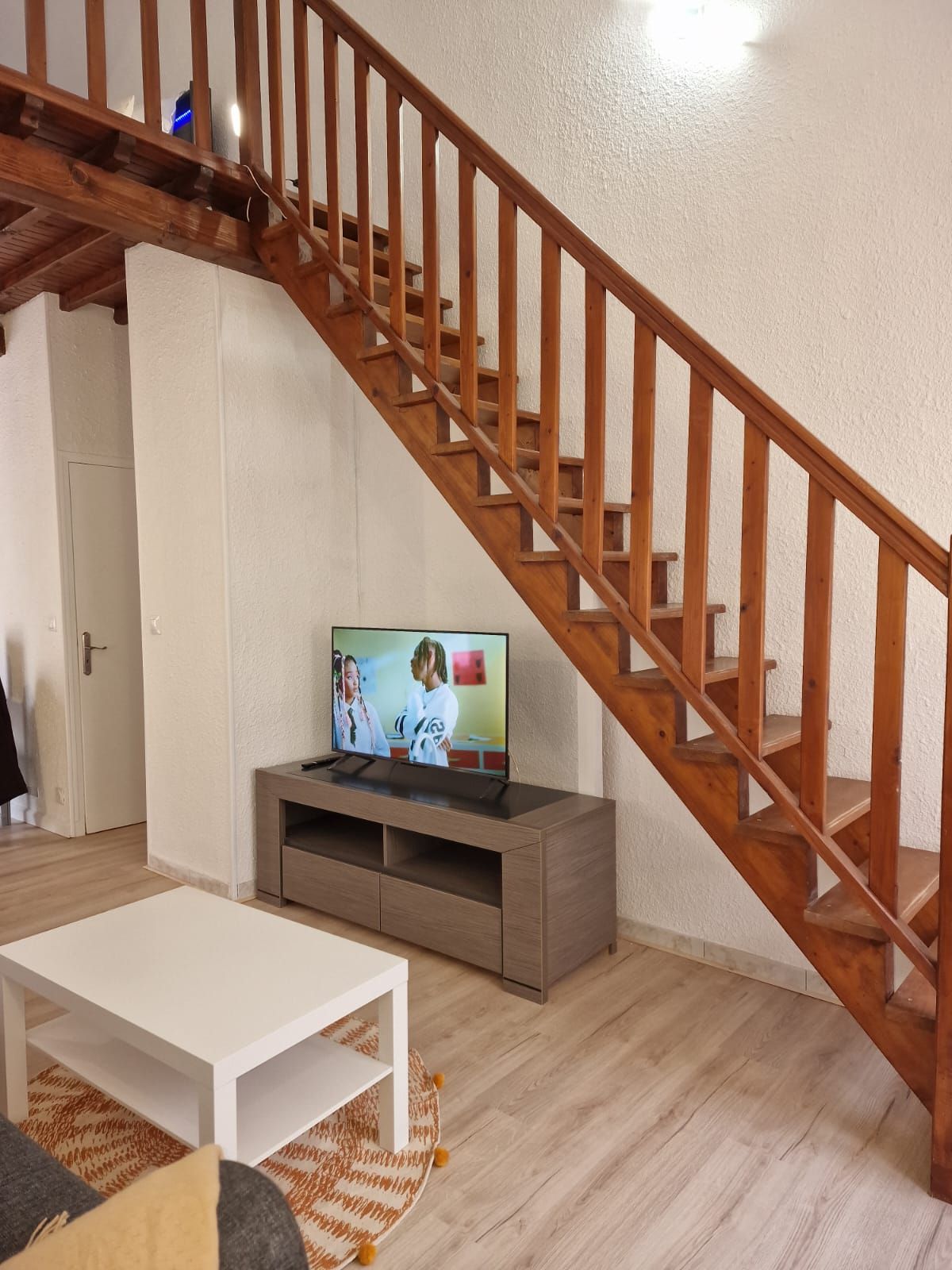 Una sala de estar con una escalera de madera, un televisor sobre un mueble de madera y una mesa de centro blanca.