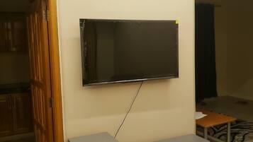Una habitación con televisión montada en la pared, un mueble con cajones y una puerta parcialmente visible.