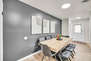 A modern dining area with a long wooden table, black chairs, and a grey accent wall with three framed artworks. - Air Hockey + Shuffleboard | 3BR Near CLE (Berea)