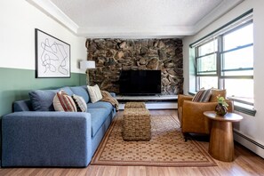 A living room with a stone wall, a blue sofa, a brown armchair, a small table, and a flat-screen TV. - Cozy 2BR, Steps to Mines, Workspaces (Golden)