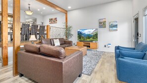 A modern living room with a brown sofa, blue armchairs, a wooden TV stand, and a large TV displaying a mountain landscape. - Deer Mountain Retreat 3bdr townhouse near Fall RiverRocky Mountain National Park (Estes Park)