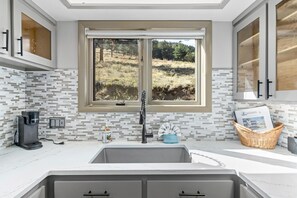 A kitchen with a window view of a grassy field, a coffee maker, and a basket with magazines. - Deer Mountain Retreat 3bdr townhouse near Fall RiverRocky Mountain National Park (Estes Park)