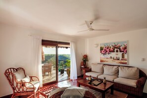 Ein Wohnzimmer mit einer Couch, einem Sessel, einem Couchtisch und einem Balkon mit Blick auf Grünflächen.