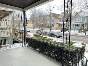 A covered porch with a metal railing, a staircase leading to a green door, and a view of a snow-covered street with houses and parked cars. - Entire Historic Elmwood Village 3BR APT - Amazing Location (Buffalo)