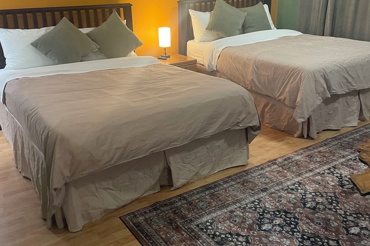 A hotel room with two beds, a nightstand with a lamp, a patterned rug, and a wooden floor.