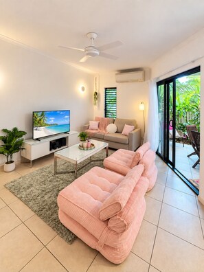 Ein modernes Wohnzimmer mit Fernseher, einer Couch und Blick auf einen tropischen Strand durch eine Glasschiebetür.