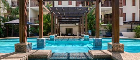 A swimming pool with a pergola structure, surrounded by a building with balconies and a clear sky.