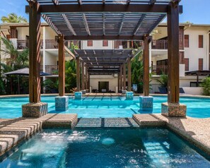 Ein Pool mit Pergola, umgeben von einem Gebäude mit Balkonen und blauem Himmel.