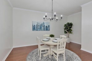 A dining room with a round table, white chairs, a chandelier, a cityscape painting, and two potted plants. - Charming, spacious, and walking distance to University of South Alabama. (Mobile)