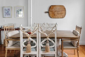 Een eetgedeelte met een houten tafel en stoelen, een witte muur met twee kunstwerken in lijsten, en een rustieke houten plank.