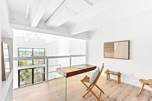 Ein geräumiges Zimmer mit hohen Decken, großen Fenstern mit Blick auf die Stadt, einem Holztisch, einem Stuhl und einem kleinen Tisch mit einem dekorativen Gegenstand.