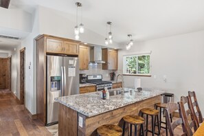A modern kitchen with a central island, stainless steel appliances, wooden cabinets, and a window with a view of greenery. - Homewood Cabin  - Walk to the Lake - Pet Friendly (Homewood)