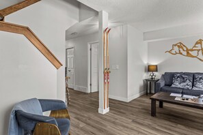 A modern living room with a sofa, a coffee table, and a lamp. There is a staircase with a wooden frame and a door leading to another room. - Mountain Retreat | 3BR | Walk to Downtown (Canmore)