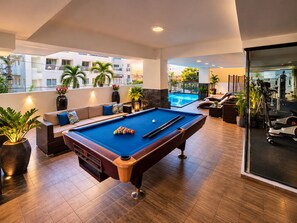 A pool table in a well-lit room with a sofa and potted plants, overlooking a swimming pool and gym area. - Modern Condo in Evaristo Morales w/Pool & Gym (Santo Domingo)