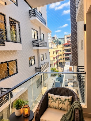A balcony with a wicker chair, cushions, and a small table with a plant and candle. - Modern Condo in Evaristo Morales w/Pool & Gym (Santo Domingo)