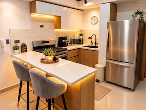 A modern kitchen with a stainless steel refrigerator, a microwave, a stove, and a sink. There are two bar stools and a small potted plant on the island. - Modern Condo in Evaristo Morales w/Pool & Gym (Santo Domingo)