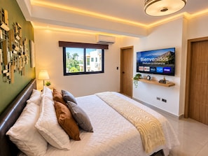 A modern hotel room with a large bed, a mounted TV displaying a welcome screen, and a window with a view of buildings. - Modern Condo in Evaristo Morales w/Pool & Gym (Santo Domingo)