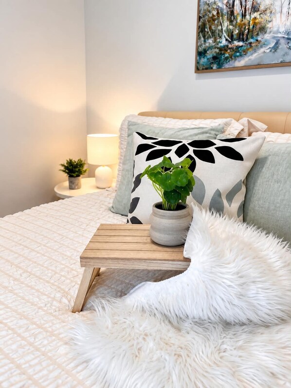 A neatly made bed with a fluffy white throw, a bedside table with a lamp, a small potted plant, and a wall-mounted painting.