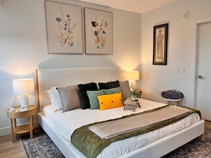 Een netjes opgemaakt bed met verschillende kussens en een deken, een nachtkastje met een lamp, en een muur met twee kunstwerken in lijsten.