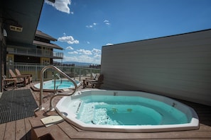 Ein Whirlpool auf einer Holzterrasse mit Liegestühlen, ein Schwimmbad und Blick auf Gebäude und Berge.