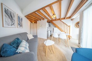 Un salon moderne avec un canapé gris, des coussins bleus, un coin repas avec des chaises blanches et un escalier menant à un niveau supérieur.