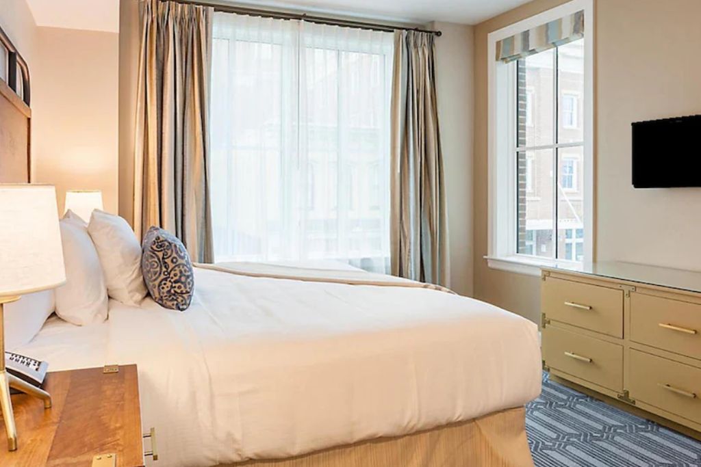 Ein Hotelzimmer mit einem Bett, einem Schreibtisch mit Lampe, einem Fenster mit Vorhängen, einem Schminktisch und einem an der Wand befestigten Flachbildfernseher.