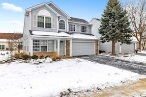 A two-story house with a garage, a snow-covered driveway, and a large evergreen tree in the front yard. - 4BR Family Home • Ideal for Insurance & Work Stays (Lake in the Hills)
