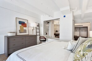 Une chambre à coucher moderne, avec un grand lit, une commode, une chaise et une salle de bain visible à travers une porte ouverte.