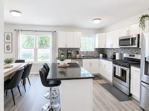 A modern kitchen with white cabinets, a black countertop island, stainless steel appliances, and a dining area with a wooden table and black chairs. - Furnished 3BR Home | Fenced Yard | Mid-Term (Grayslake)