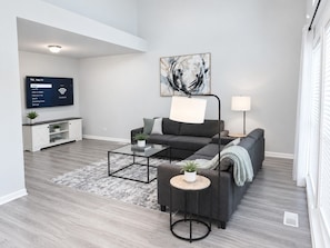 A modern living room with a grey sofa, a coffee table, and a TV mounted on the wall. - Furnished 3BR Home | Fenced Yard | Mid-Term (Grayslake)