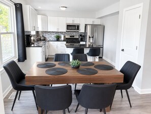 Eine moderne Küche mit weißen Schränken, Edelstahlgeräten und einem Essbereich mit Holztisch und schwarzen Stühlen.