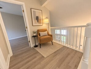 A room with a wooden floor, a white railing, a chair, a lamp, and a framed picture on the wall. - Furnished 3BR Home | Fenced Yard | Mid-Term (Grayslake)