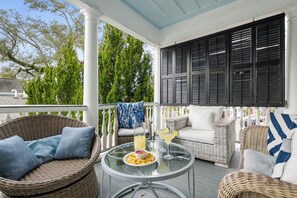 Eine überdachte Veranda mit Korbmöbeln, einem Glastisch und Blick auf Bäume und eine Straße.