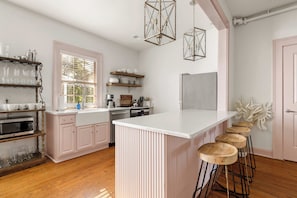 A kitchen with a central island, wooden bar stools, and a window with blinds. - The Waltz | 4 BR Boho Retreat | Walk to King (Charleston)