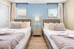 Habitación de hotel con dos camas, una mesita de noche con lámpara y vistas al exterior a través de las ventanas.