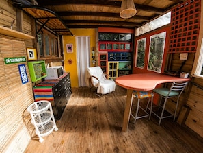 A rustic wooden room with a dining table, chairs, a white chair, a small white stool, and a shelf with various items. - Cabana Caapora: tranquilidade em meio à natureza em Florianópolis (Florianópolis)