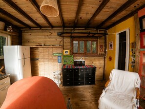 A rustic cabin interior with a wooden ceiling, a white refrigerator, a black storage unit, and a white chair. - Cabana Caapora: tranquilidade em meio à natureza em Florianópolis (Florianópolis)