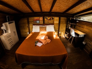A wooden cabin room with a bed, a desk, and a fan.