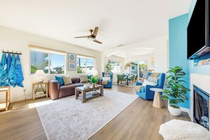 Een ruime woonkamer met een bruine leren bank, blauwe fauteuils, een salontafel en een plafondventilator.