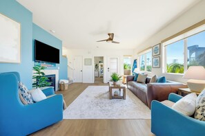 A spacious living room with a large TV, blue sofas, a coffee table, and a view of the outdoors through large windows. - La Jolla Beach House -  Walk to Windansea, EV Charger (San Diego)