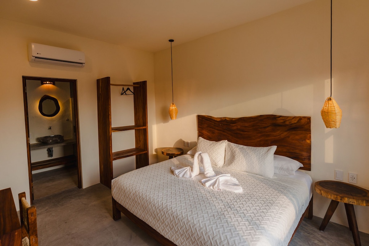 A bedroom with a large bed, wooden headboard, bedside tables, a hanging light, and an air conditioning unit.