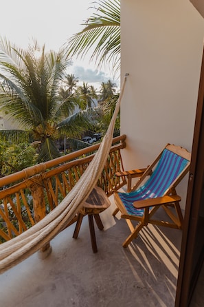 A balcony with a hammock, wooden chairs, and a view of palm trees and a beach.