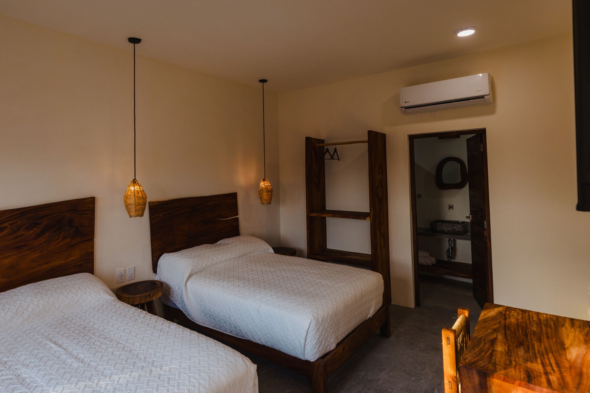 Ein Hotelzimmer mit zwei Betten, einem Holz-Kopfteil, einem kleinen Nachttisch, einer Pendelleuchte und einer Klimaanlage.