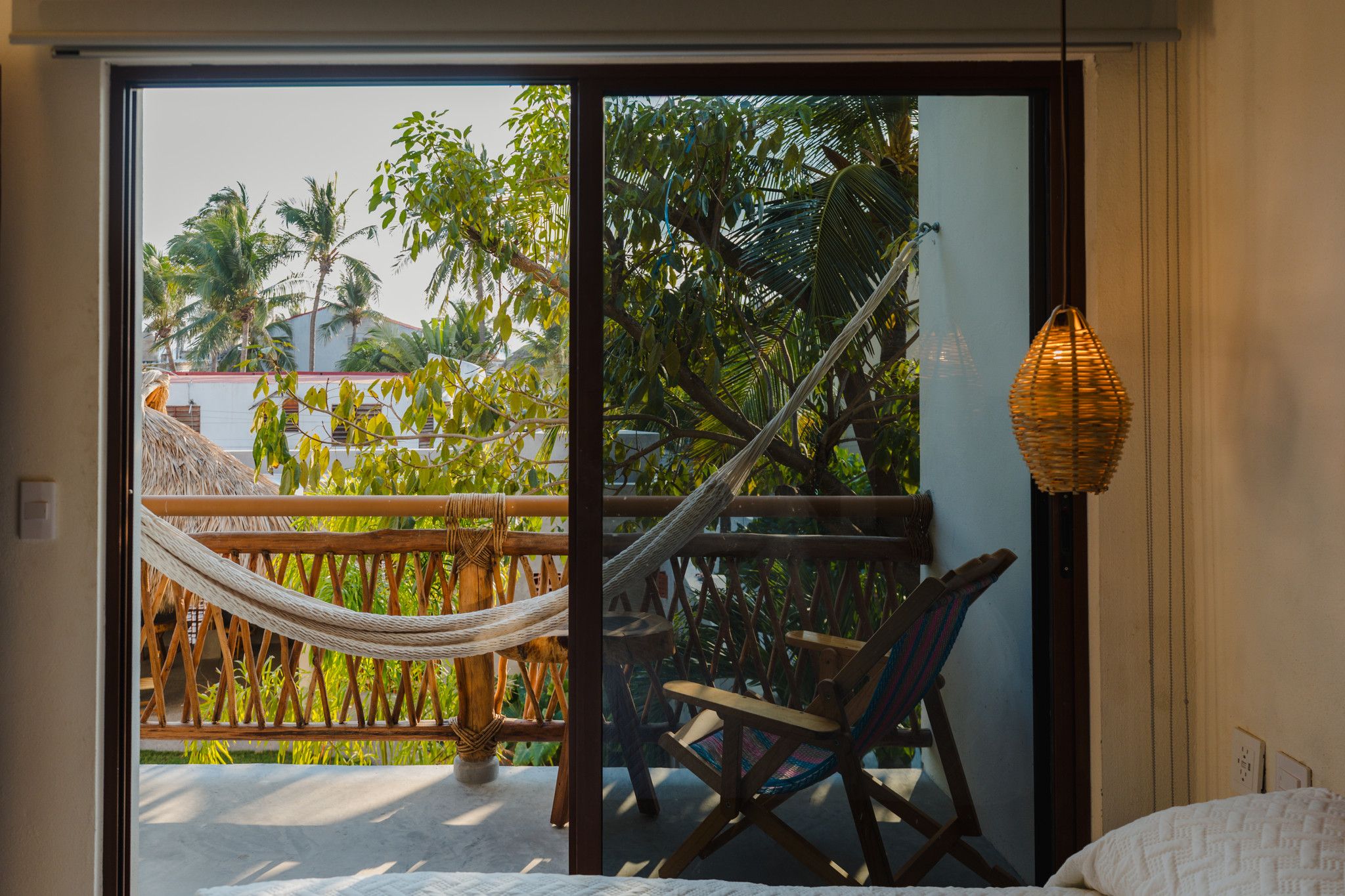 A room with a bed, a hammock, a chair, and a view of palm trees through a glass door.
