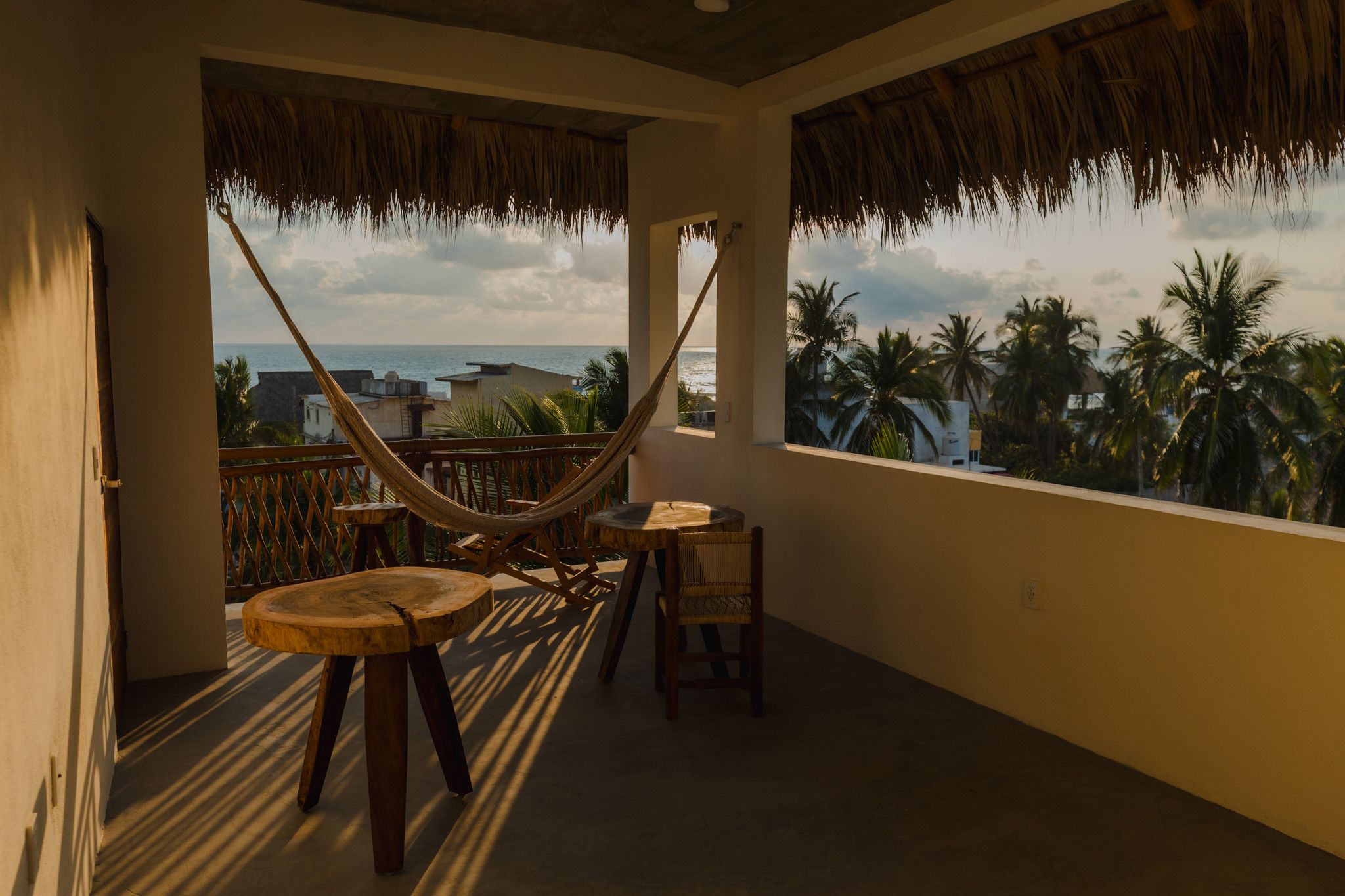 A balcony with a hammock, wooden furniture, and a view of the ocean and palm trees.