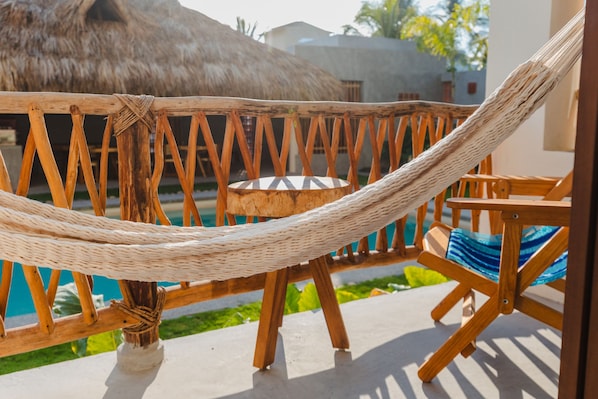 Una hamaca atada entre postes de madera, con vista a un área de piscina con una estructura de techo de paja y una mesa de madera con cojín azul.