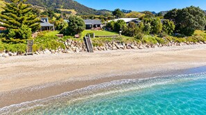 A beachfront property with a clear view of the ocean, a stone pathway leading to the beach, and a well-maintained lawn with a building in the background. - Pakawau Beach - Ratrace Retreat (Pākawau)