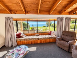 A room with a wooden ceiling, a large window with a view of the sea, a cushioned bench with pillows, and a brown armchair. - Pakawau Beach - Ratrace Retreat (Pākawau)