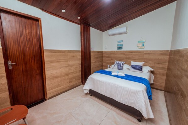 A hotel room with a wooden door, a bed with a blue blanket, a white bedspread, and a wall-mounted air conditioner.