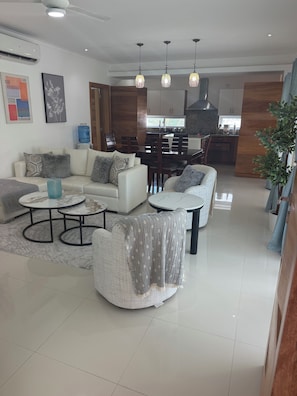 A modern living room with a sofa, armchairs, and a coffee table. There is a dining area with a table and chairs, and a kitchen in the background. - An exquisite an exceptional, 5 star resorts style condominium complex (Punta Cana)